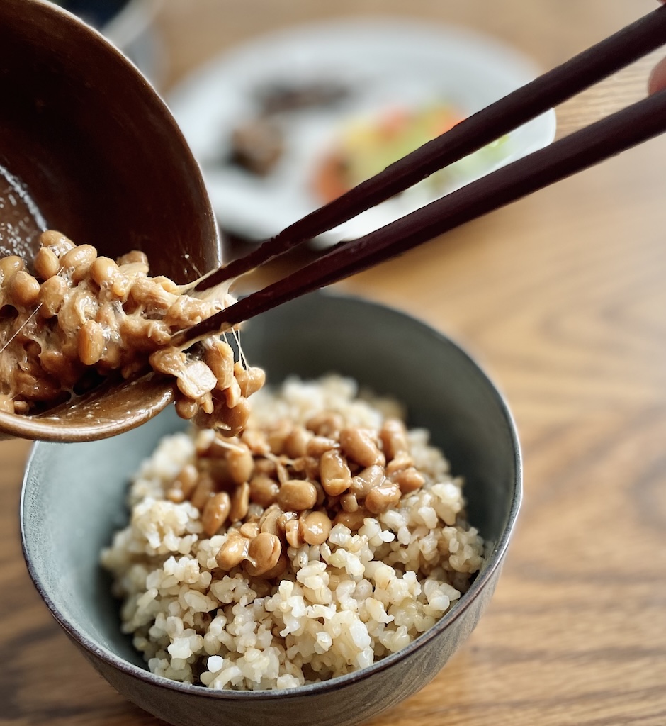 A close-up shot of chopsticks lifting natto from a bowl, with the natto on top of a serving of rice in a gray bowl.